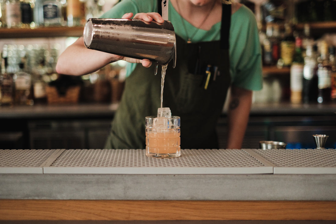 Bartender schenkt een cocktail uit een shaker in een glas achter een bar met flessen en barmateriaal