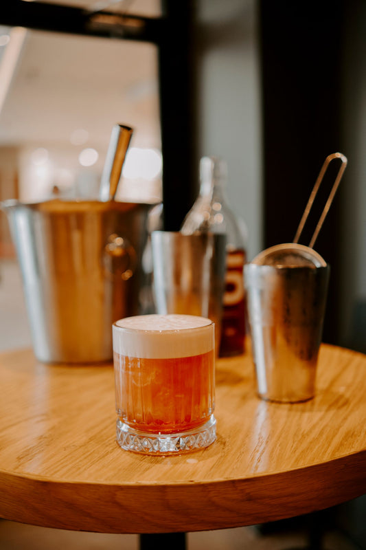 Amaretto Sour cocktail met schuimlaag en sinaasappelgarnering op een bar naast cocktailtools
