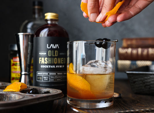Citroenschil wordt boven een tumbler met amberkleurige whisky uitgeknepen, zoals bij het afwerken van een Sazerac cocktail