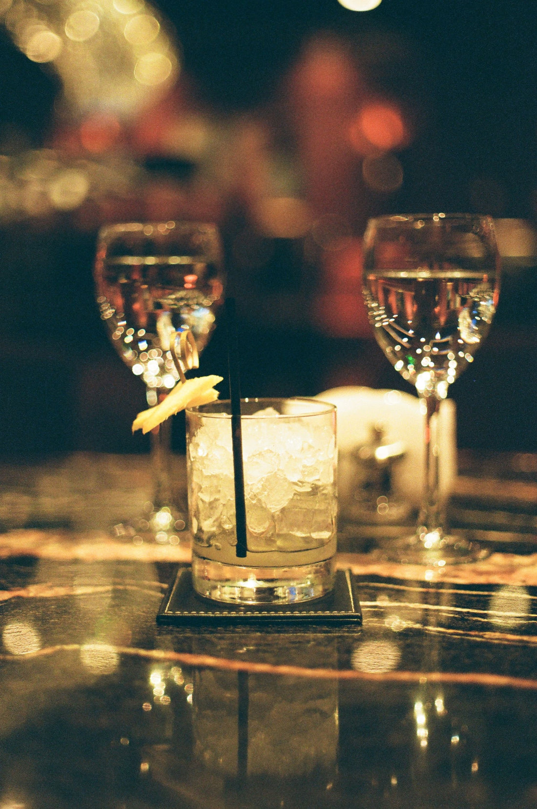 Cocktail met ijsblokjes en een schijfje citroen op een spiegelende bar, met wijnglazen op de achtergrond